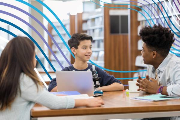 Three students sit around a table in a library or classroom engaged in conversation and learning. A student in the center smiles while speaking to another student across the table. A laptop, microscope, notebooks, and school supplies are on the table. Blue curved graphic lines overlay the image.
